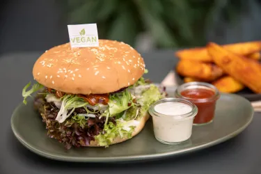 Ein leckerer veganer Burger mit frischem Salat und einer Sesambrötchen. Dazu gibt es zwei Dips und knusprige Süßkartoffelpommes.