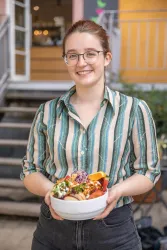 Eine lächelnde junge Frau hält eine Schüssel mit buntem Essen. Im Hintergrund sind Stufen und ein Gebäude sichtbar.