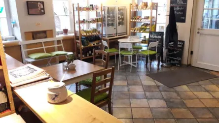 Ein gemütliches Café mit Holztischen und bunten Stühlen. Im Hintergrund sind Regale mit verschiedenen Produkten und Getränken zu sehen.