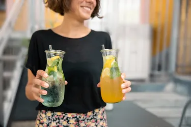 Eine Frau hält zwei Getränke in Glasbehältern. Eines ist klar mit Zitrone und Minze, das andere ist orange.
