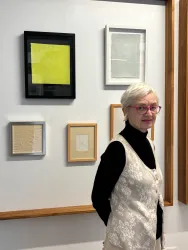 Eine ältere Frau mit einem eleganten Look steht vor einer Wand mit mehreren gerahmten Kunstwerken. Die Bilder variieren in Form und Farbe, darunter ein leuchtend gelbes.