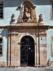 Eine beeindruckende Eingangstür mit kunstvollem Steinmetzwerk und einem Skulpturenrelief über dem Tor. Die Fassade ist in sanften Farben gehalten und hat Fenster auf beiden Seiten.