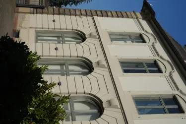 Ein modernes Gebäude mit geschwungenen Fenstern und einem klaren Design. Das Bild zeigt eine helle Fassade, umgeben von Pflanzen.