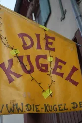 Ein großes, gelbes Schild mit der Aufschrift "DIE KUGEL". Es enthält auch eine Webadresse am unteren Rand.