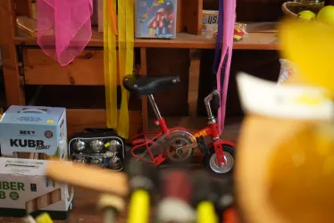 Ein kleines rotes Spielzeugfahrrad steht in einem Ladenregal. Umgeben von bunten Bändern und verschiedenen Gegenständen.