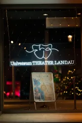 Ein beleuchtetes Schaufenster des Universum Theaters in Landau. Vor dem Fenster steht ein Plakat für eine bevorstehende Aufführung.