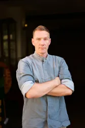 Ein Mann steht mit verschränkten Armen und ernstem Blick in einem Innenraum. Er trägt ein hellblaues Hemd und wirkt selbstbewusst.