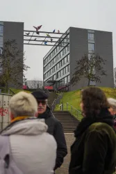 Ein modernes Gebäude mit bunten Vögeln auf einer Brücke im Vordergrund. Eine Gruppe von Menschen steht davor und scheint zu beobachten.