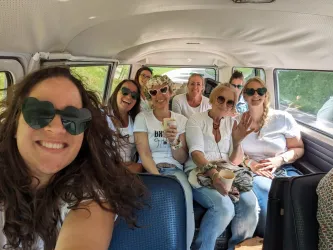Eine Gruppe von fröhlichen Frauen sitzt in einem Fahrzeug und lächelt in die Kamera. Sie tragen Sonnenbrillen und haben Getränke in den Händen.