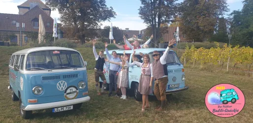 Zwei blaue Volkswagen-Busse stehen auf einer Wiese, umgeben von Bäumen und Weinreben. Eine Gruppe von fünf Personen posiert fröhlich neben den Autos.