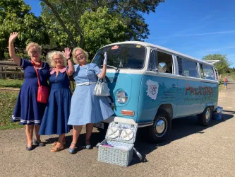 Drei Frauen in eleganten Kleidern stehen fröhlich vor einem blauen VW-Bus. Sie winken in die Kamera und genießen einen sonnigen Tag.