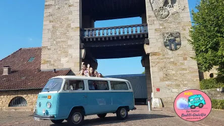 Ein blauer VW-Bully steht vor dem Deutschen Weintor. Eine Gruppe von Personen sitzt fröhlich im oder auf dem Fahrzeug.