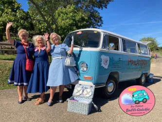 Drei Frauen in stylischen Kleidern stehen fröhlich neben einem bunten Oldtimer-Bus. Im Hintergrund ist eine grüne Landschaft sichtbar.