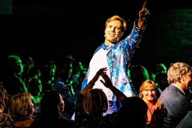 Ein Sänger in einem glitzernden Anzug sorgt für gute Stimmung auf der Bühne. Das Publikum ist begeistert und klatscht mit.