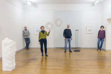 In einer Galerie stehen vier Personen verteilt im Raum. Es gibt verschiedene Kunstwerke, darunter eine weiße Skulptur und Zeichnungen an den Wänden.