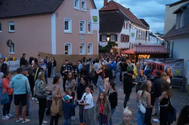 Eine lebhafte Straßenszene mit vielen Menschen, die sich versammeln. Die Umgebung ist von bunten Gebäuden und stimmungsvoller Beleuchtung umgeben.