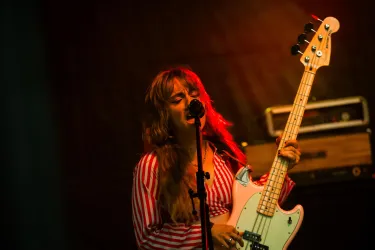 Eine Musikerin spielt Bass und singt auf einer Bühne. Sie trägt ein gestreiftes Oberteil und hat lange, lockige Haare.