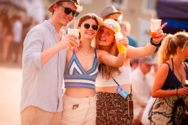 Drei junge Menschen lächeln und stoßen mit Getränken an. Sie tragen Sonnenbrillen und sommerliche Kleidung auf einem fröhlichen Fest.
