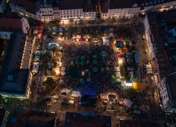 Eine belebte Platzansicht mit vielen Menschen und bunten Ständen. Die Atmosphäre ist festlich und bunt, ideal für ein Event oder Festival.