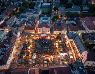 Ein lebhaftes Stadtfest mit einer großen Menschenmenge in einem zentralen Platz. Bunte Lichter und Stände schaffen eine festliche Atmosphäre.