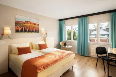 Ein helles Schlafzimmer mit einem Doppelbett und orangefarbenen Kissen. Große Fenster mit Vorhängen und ein kleiner Tisch in der Ecke ergänzen den Raum.