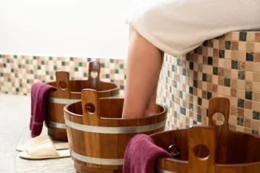 Eine entspannende Wellness-Szene mit hölzernen Fußbädern. Ein Bein wird in eines der Bäder eingetaucht, daneben liegen Handtücher.