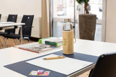 Ein moderner Besprechungsraum mit einem weißen Tisch und schwarzen Stühlen. Auf dem Tisch liegen Schreibmaterialien und eine Wasserflasche.