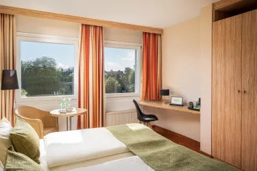 Ein modernes Hotelzimmer mit zwei Betten und einem Schreibtisch. Große Fenster bieten viel natürliches Licht und einen Blick ins Grüne.