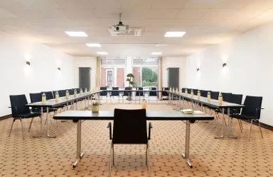 Ein moderner Konferenzraum mit einem großen Tisch und Stühlen in U-Form. Der Raum ist gut beleuchtet und hat Fenster, die Tageslicht hereinlassen.