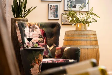 Ein gemütlicher Sessel mit buntem Stoff steht neben einem Holzfass. Im Hintergrund sind mehrere Bilder an der Wand zu sehen.