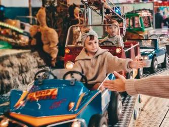Ein Kind sitzt in einem kleinen Spielzeugauto und lächelt. Im Hintergrund sind weitere Autos und festliche Lichter zu sehen.