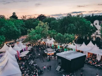 Eine belebte Veranstaltung im Freien mit vielen Festzelten und Bäumen. Im Hintergrund ist eine Bühne zu sehen, während der Himmel sanfte Farben zeigt.