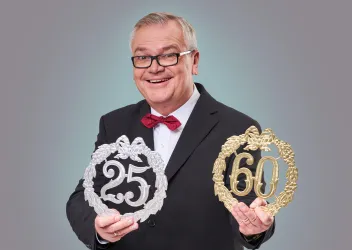 Ein fröhlicher Mann in einem Anzug hält zwei dekorative Kränze in den Händen. Einer ist silber und steht für 25 Jahre, der andere ist gold und symbolisiert 60 Jahre.