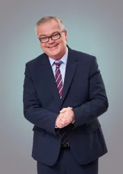 Ein lächelnder Mann in einem Anzug steht mit gefalteten Händen. Der Hintergrund ist ein sanftes Grau.