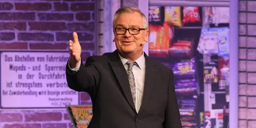 Ein Mann trägt einen Anzug und steht auf einer Bühne. Er lächelt und spricht in ein Mikrofon, während im Hintergrund verschiedene Snacks und Getränke zu sehen sind.