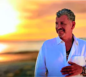 Ein Mann mit einem weißen Hemd steht am Strand und lächelt. Im Hintergrund ist ein schöner Sonnenuntergang zu sehen.