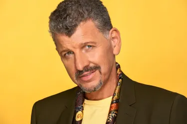 Ein Mann mit grauen Locken und einem Schnurrbart lächelt in die Kamera. Er trägt eine schwarze Jacke und ein gelbes T-Shirt vor einem gelben Hintergrund.