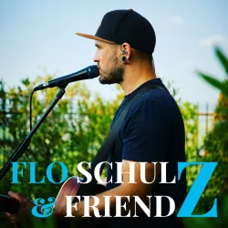 Ein Musiker steht mit einer Gitarre auf der Bühne und singt in ein Mikrofon. Im Hintergrund sind grüne Pflanzen zu sehen.