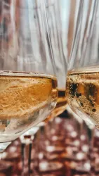 Zwei Gläser Sekt mit sprudelndem Inhalt. Im Hintergrund sind unscharfe Gläser und eine festliche Atmosphäre zu sehen.