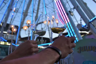 Zwei Gläser mit Wein werden hochgehalten, während im Hintergrund ein Riesenrad mit bunten Lichtern leuchtet. Die Atmosphäre ist feierlich und festlich.