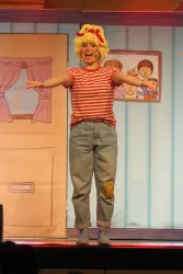 Ein fröhliches Kind auf der Bühne trägt ein gestreiftes Shirt und eine gelbe Perücke. Im Hintergrund sind Bilder und ein Fenster zu sehen.