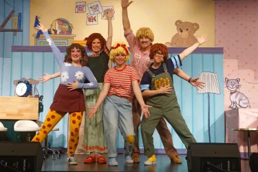 Eine Gruppe von fünf Personen in bunten Kostümen steht auf einer Bühne. Sie posieren fröhlich vor einer kindgerechten Kulisse.