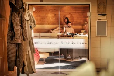 Eine finnische Sauna mit Holzverkleidung und entspannenden Geräten. Im Hintergrund sind zwei Personen sichtbar, die sich ausruhen.