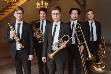 Eine Gruppe von fünf Musikern in Anzügen steht zusammen und hält verschiedene Blechblasinstrumente. Sie posieren in einem eleganten Raum mit Kronleuchtern im Hintergrund.