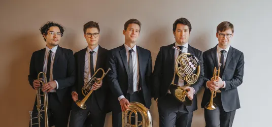 Eine Gruppe von fünf Musikern in Anzügen steht aufgereiht zusammen. Jeder hält ein Blasinstrument in der Hand.