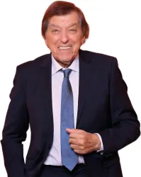Ein älterer Mann lächelt freundlich in einem dunklen Anzug mit einer blauen Krawatte. Er steht leger mit einer Hand an seinem Anzug.