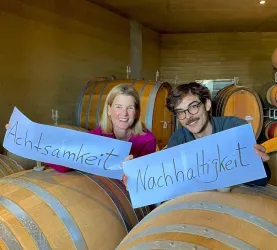 Zwei Personen stehen in einem Weinkeller und halten Plakate mit den Wörtern „Achtsamkeit“ und „Nachhaltigkeit“. Im Hintergrund sind Weinfässer zu sehen.
