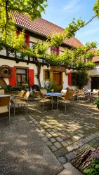 Ein einladender Innenhof mit Tischen und Stühlen, umgeben von blühenden Pflanzen. Im Hintergrund sieht man ein traditionelles Haus mit roten Fensterläden.