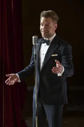Ein Sänger in einem eleganten Anzug steht vor einem Mikrophon. Er lächelt und scheint bereit zu performen.
