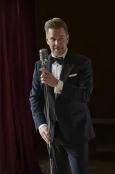 Ein Mann in einem eleganten Anzug und einer Fliege steht vor einem Mikrofon. Der Hintergrund ist dunkelrot und schafft eine festliche Atmosphäre.